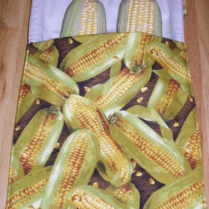 May include: A white fabric bag with a brown background and a pattern of corn cobs. Two ears of corn are inside the bag.