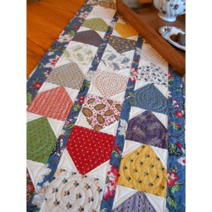 May include: A blue and white table runner with a patchwork design featuring various floral patterns and a swirl design. The runner is made of fabric and has a quilted texture.