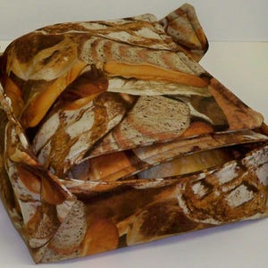Puede incluir: Una cesta de pan de tela marrón y beige con un patrón de rebanadas de pan. La cesta está doblada y tiene una forma cuadrada.
