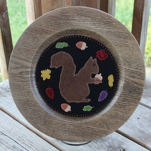 May include: A wooden plate with a felt appliqué of a brown squirrel holding an acorn. The squirrel is surrounded by felt leaves and acorns in various colors, including red, green, yellow, and purple. The plate is a decorative piece for a home.