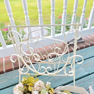 May include: Off-white metal magazine rack with decorative scrollwork. The rack is on a weathered teal surface, with a bouquet of flowers in front. The background includes a white porch railing and green grass.