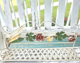 May include: A cream-colored metal wall shelf featuring a lattice-patterned base and a decorative top rail. The rail is adorned with painted metal accents, including green leaves, a pale yellow leaf, and two small, muted pink rose-shaped flowers.