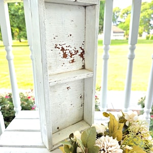 May include: A tall, rectangular wooden box with a distressed white painted finish, featuring a single horizontal shelf in the middle. The wood shows signs of wear with brown patches visible through the chipped white paint on the interior back panel.