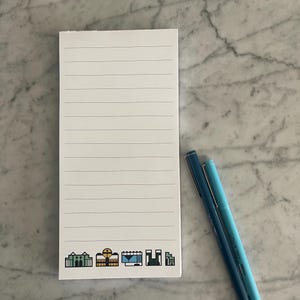 Op de afbeelding: Een wit notitieboekje met gestreept papier en een rij kleurrijke stadssiluet-pictogrammen aan de onderkant. Twee blauwe pennen bevinden zich aan de rechterkant van het notitieboekje.