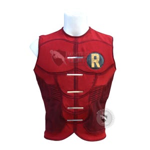 May include: A sleeveless red vest featuring a textured fabric, a round neckline, and four horizontal silver-toned metallic bars across the chest. A circular black patch with a gold letter R is positioned on the upper left side of the chest.