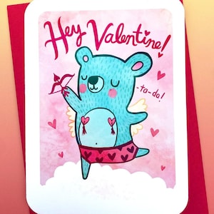 Puede incluir: Una tarjeta de San Valentín con un oso azul con alas, sosteniendo un arco y una flecha, y usando ropa interior rosa con corazones. La tarjeta tiene un fondo rosa acuarela y el texto "Hey Valentine! -ta-da!"