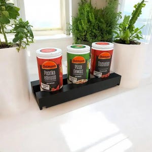 Könnte beinhalten: Drei Gewürzgläser mit roten, grünen und weißen Etiketten stehen auf einem schwarzen Gewürzregal aus Holz. Die Gläser sind mit "Paprika edelsüß", "Pizza Gewürz" und "Tomaten" beschriftet.