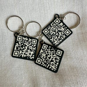 Puede incluir: Tres llaveros cuadrados negros con un diseño de código QR y anillos plateados. Los llaveros están colocados sobre una superficie de tela blanca. Los códigos QR son blancos sobre un fondo negro.