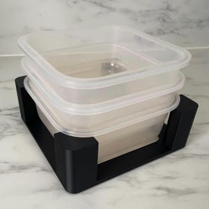 Può includere: Due contenitori per alimenti in plastica trasparente con coperchi, impilati su un supporto quadrato nero. I contenitori sono rettangolari e vuoti. Il supporto ha un design minimalista.