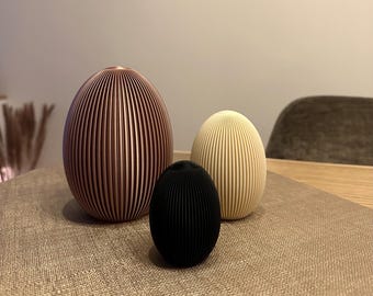 May include: Three decorative egg-shaped objects in varying sizes and colors. The largest is a rose gold color, the medium-sized one is cream, and the smallest is black. All have a ribbed, vertical design. They are arranged on a neutral-toned surface.