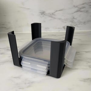 Può includere: Un set di contenitori per alimenti in plastica nera con coperchi trasparenti e un supporto nero. Il set include più contenitori con coperchi trasparenti a scatto. I contenitori sono impilati.