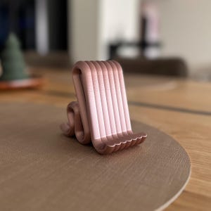 Könnte beinhalten: Ein roségoldener Telefonständer mit einem wellenförmigen, vertikalen Design. Der Ständer hat eine Reihe paralleler, gebogener Linien, die eine Halterung für ein Telefon oder Tablet bilden. Die Basis ist eine Reihe kleiner, gebogener Bögen.