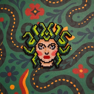 Könnte beinhalten: Eine Medusa-Kopf-Anstecknadel aus Perlen mit grünen, schwarzen und beigefarbenen Perlen. Die Anstecknadel zeigt ein detailliertes Gesicht mit roten Lippen und einem grünen, schlangenartigen Haardesign. Der Hintergrund ist ein grüner Stoff mit einem sich wiederholenden Muster aus sich windenden schwarzen Linien und roten Blumen.
