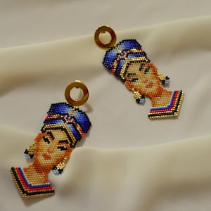 Pode incluir: Um par de brincos de contas com um retrato colorido de uma mulher com um turbante azul. Os brincos são feitos com contas pequenas e têm argolas douradas.