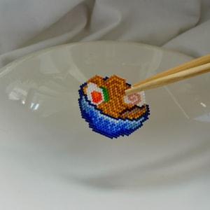 Puede incluir: Un rollo de sushi de cuentas con detalles naranjas, blancos, verdes y rosas, sostenido por palillos de madera. El sushi está en un cuenco de cuentas azul, presentado en un plato blanco. El sushi está hecho de pequeñas cuentas de colores.