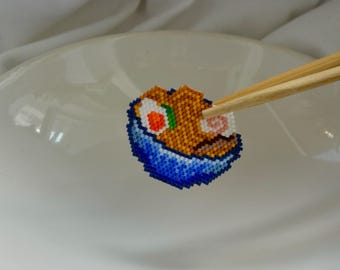 Puede incluir: Un rollo de sushi de cuentas con detalles naranjas, blancos, verdes y rosas, sostenido por palillos de madera. El sushi está en un cuenco de cuentas azul, presentado en un plato blanco. El sushi está hecho de pequeñas cuentas de colores.