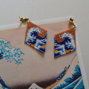Könnte beinhalten: Diamantförmige Ohrringe mit einem pixeligen Wellenmuster in Blau, Weiß und Gelb, vor einem rostfarbenen Hintergrund. Die Ohrringe haben goldfarbene Pfosten. Inspiriert von japanischer Kunst.