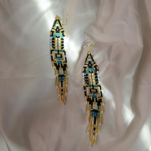 May include: A pair of beaded earrings with a geometric design. The earrings feature a pattern of black, white, and turquoise beads, accented with gold-colored beads. The earrings have a long, dangling design.