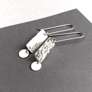 May include: A pair of silver-toned drop earrings featuring a rectangular pendant with a hammered, textured surface. Each rectangular piece is attached to a small, smooth circular charm at the bottom and hangs from a long, curved ear wire hook.