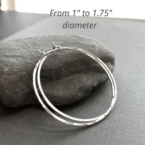 May include: A pair of silver hoop earrings with a thin, circular wire design. The earrings feature a textured surface and have a diameter ranging from 1 inch to 1.75 inches as indicated by the text in the image.