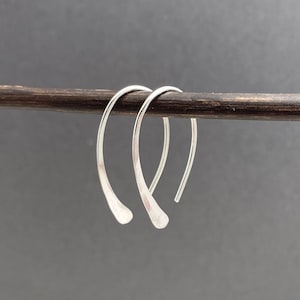 May include: A pair of silver hoop earrings with a hammered texture. The earrings are curved and have a teardrop shape at the bottom. They are displayed on a dark brown wooden rod against a gray background.