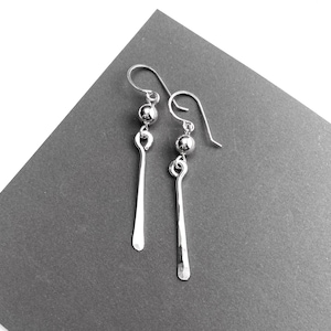 May include: A pair of silver-toned drop earrings featuring a polished spherical bead attached to a long, slender, hammered metal bar. The earrings are suspended from curved ear wires, showcasing a reflective metallic finish and a simple, elongated design.
