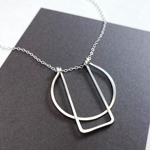 May include: A silver necklace with a geometric pendant featuring a circle and a rectangle. The pendant is made of thin metal wire and hangs from a delicate silver chain.