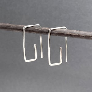 May include: A pair of silver-colored, square-shaped earrings. Each earring has a rectangular design with a small, open hook at the bottom. The earrings are displayed on a dark brown, cylindrical object against a gray background.