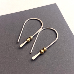 May include: A pair of silver-toned drop earrings with a curved design. Each earring features two small, gold-colored beads and a flat, rectangular end. The earrings are displayed on a dark gray surface.
