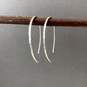 May include: A pair of silver-colored, curved earrings. The earrings are displayed on a wooden rod against a gray background. The earrings have a hammered texture on the curved portion and a smooth, straight post.