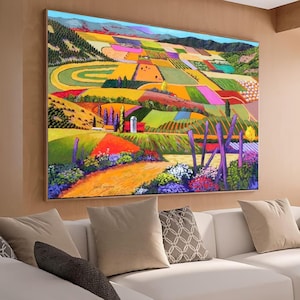 Könnte beinhalten: Ein großes, gerahmtes Landschaftsgemälde, das ein farbenfrohes Patchwork aus Feldern und Hügeln zeigt. Das Kunstwerk zeigt leuchtende Farben von Grün, Gelb, Orange und Lila, mit einem Feldweg, der zu einem Bauernhof führt. Das Gemälde wird über einem weißen Sofa ausgestellt.