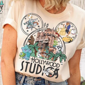 Peut inclure: Un t-shirt beige avec un imprimé graphique d'oreilles de Mickey Mouse. Les oreilles sont remplies de divers personnages Disney, dont Belle et la Bête, Woody et Buzz l'Éclair, et la Tour de la Terreur. Le texte "HOLLYWOOD STUDIOS" est imprimé sous les oreilles.