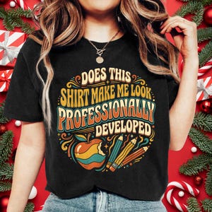 Könnte beinhalten: Ein schwarzes T-Shirt mit einer bunten Grafik und dem Text "DOES THIS SHIRT MAKE ME LOOK PROFESSIONALLY DEVELOPED" in einer Retro-Schriftart. Das Design zeigt einen angebissenen Apfel und zwei Stifte in Orange, Gelb, Blaugrün und Creme.