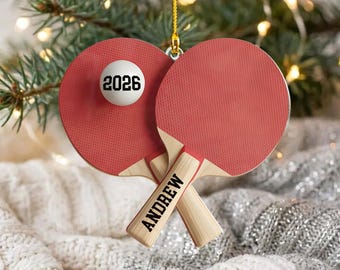 May include: A decorative ornament featuring two red table tennis paddles crossed over each other. One paddle handle is printed with the name ANDREW in black text. A white ping pong ball is attached, featuring the black printed year 2026.