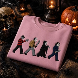 Peut inclure: Un sweat-shirt à col rond rose clair avec une broderie représentant quatre personnages de films d'horreur emblématiques marchant à la file indienne. Les personnages incluent une silhouette en pull rayé rouge et noir, un porteur de masque de hockey.