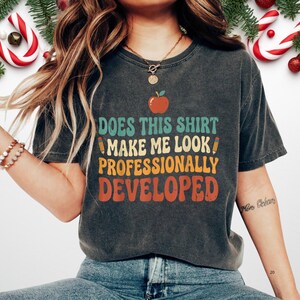 May include: A charcoal grey short-sleeve t-shirt featuring a red apple graphic, two yellow pencils, and the text "DOES THIS SHIRT MAKE ME LOOK PROFESSIONALLY DEVELOPED" in stacked, multi-coloured retro-style lettering across the chest area.