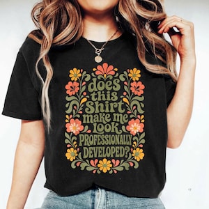 Peut inclure: Un t-shirt noir à manches courtes arborant un motif central avec le texte "does this SHIRT make me LOOK PROFESSIONALLY DEVELOPED?" entouré d'une bordure décorative composée de fleurs stylisées et de vignes feuillues orange, jaunes et vertes.