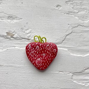 May include: A heart-shaped, red fabric item with a white strawberry pattern and green yarn detail. The item is likely a decorative pin or patch, with a textured appearance and a whimsical design.