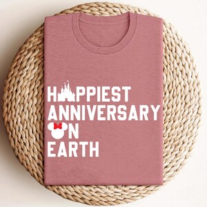 May include: A dusky pink t-shirt featuring the white text "Happiest Anniversary on Earth". The design includes a castle silhouette and a Minnie Mouse bow. The shirt is displayed on a woven, circular mat.
