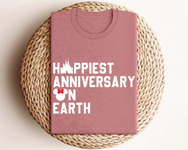 May include: A dusky pink t-shirt featuring the white text "Happiest Anniversary on Earth". The design includes a castle silhouette and a Minnie Mouse bow. The shirt is displayed on a woven, circular mat.