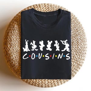K&ouml;nnte beinhalten: Schwarzes T-Shirt mit Zeichentrickfiguren und dem Wort "COUSINS" in Wei&szlig;, mit farbigen Punkten. Das Shirt wird auf einer gewebten, kreisf&ouml;rmigen Oberfl&auml;che pr&auml;sentiert.