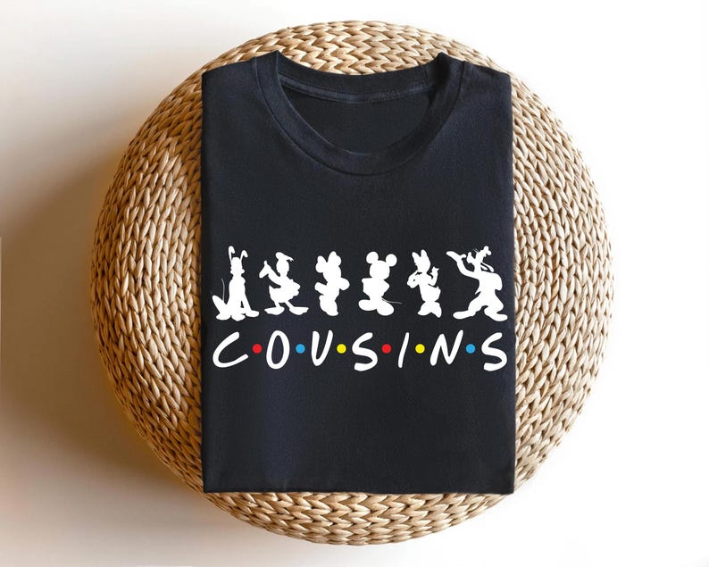 K&ouml;nnte beinhalten: Schwarzes T-Shirt mit Zeichentrickfiguren und dem Wort "COUSINS" in Wei&szlig;, mit farbigen Punkten. Das Shirt wird auf einer gewebten, kreisf&ouml;rmigen Oberfl&auml;che pr&auml;sentiert.