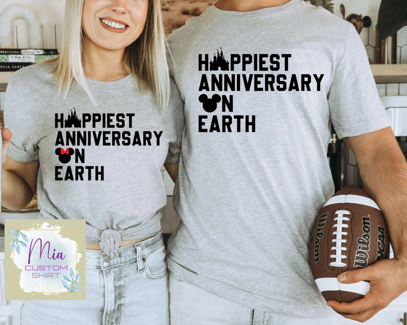 May include: Two light grey t-shirts with the text "HAPPIEST ANNIVERSARY ON EARTH" in black, featuring a castle and Mickey Mouse head graphic. One shirt has a red bow. The shirts are for couples. A football is also visible.