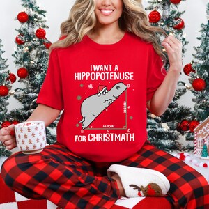 May include: Red t-shirt with the text "I want a Hippopotanuse for Chrismath" and a cartoon hippo wearing a Santa hat. The shirt is paired with red and black plaid pajama pants. A white mug with a festive design is held in the hand.