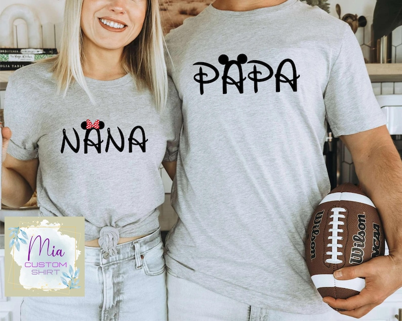 May include: Two grey t-shirts with a Disney-inspired design. The shirt on the left says "NANA" with a red bow and the shirt on the right says "PAPA" with Mickey Mouse ears.