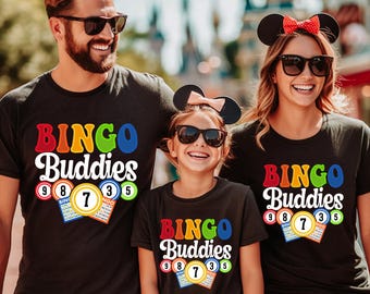 May include: Black t-shirts with the text "BINGO Buddies" in colorful letters. Each shirt features bingo balls and cards with numbers. The image shows a family wearing matching shirts, suggesting a family outing or event.