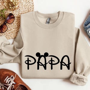 May include: A tan sweatshirt with a black graphic of Mickey Mouse ears and the word "PAPA" in a stylised font.