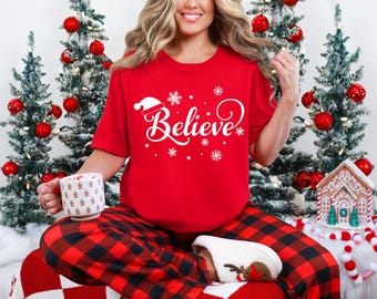 May include: A red t-shirt with the word "Believe" in white script, a Santa hat, and snowflakes. The person is wearing red and black plaid pajama pants and holding a mug. Christmas trees and decorations are in the background.