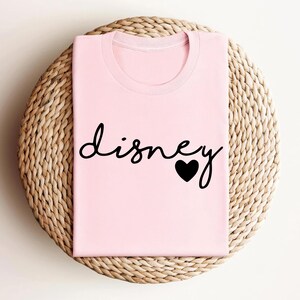 May include: A light pink t-shirt featuring the word "disney" written in a black cursive script font, with a solid black heart icon replacing the dot on the letter i. The garment has a classic crew neckline and short sleeves, presented folded.