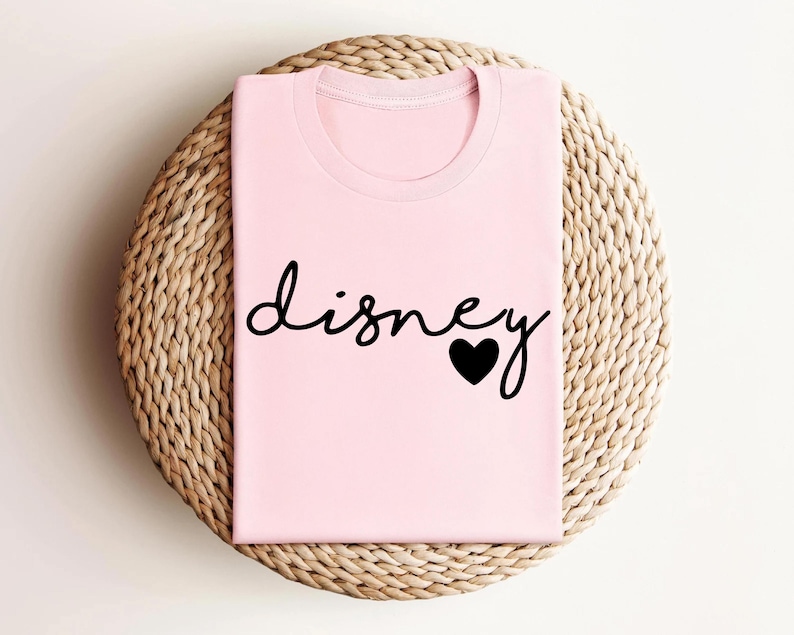 May include: A light pink t-shirt featuring the word "disney" written in a black cursive script font, with a solid black heart icon replacing the dot on the letter i. The garment has a classic crew neckline and short sleeves, presented folded.